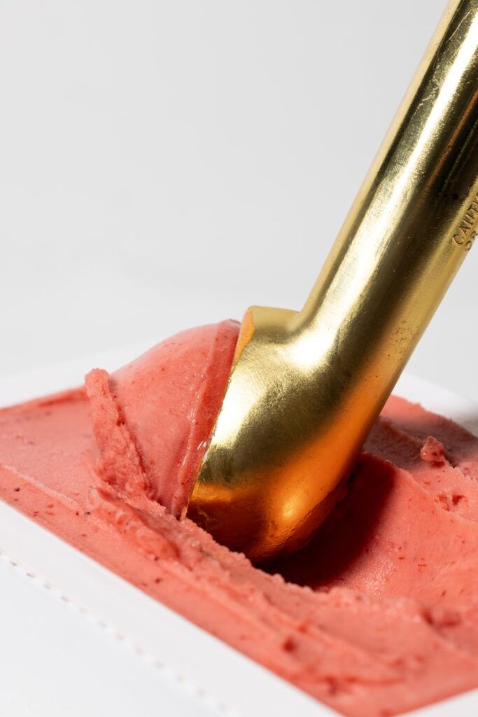 Person scooping pink gelato with gold spoon.