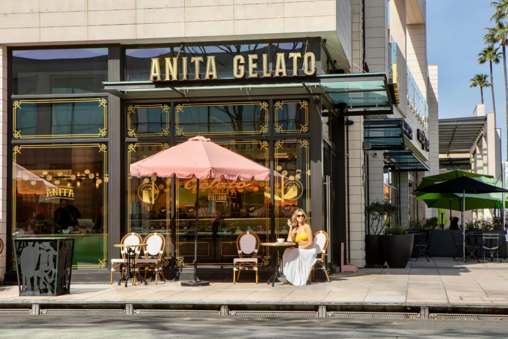 people sitting at tables outside a restaurant with umbrellas on the sidewalk