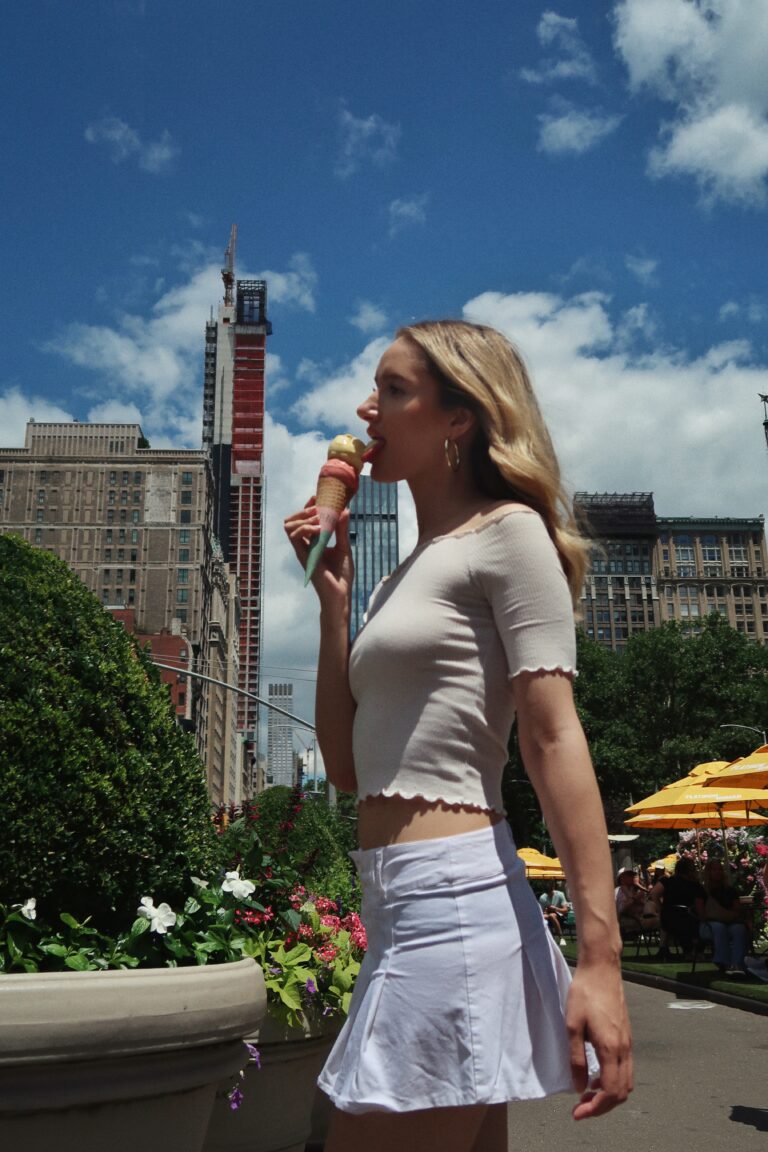 Blond woman in white skirt enjoying ice cream cone in city.