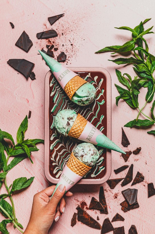 a person holding two ice cream cones in a container with chocolate and mint leaves